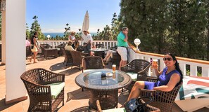 Relax on the ocean-view patio with wicker furniture and a glass table, surrounded by trees and umbrellas for shade.