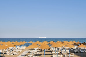 A beach with rows of sun loungers and thatched umbrellas, a cruise ship in the distance, and a clear blue sky.