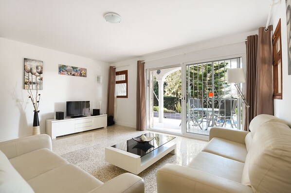 A modern living room with a sofa, a coffee table, a TV, and a sliding glass door leading to an outdoor area with a table and chairs.