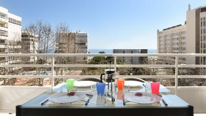 Un balcón con una mesa lista para dos personas, con vistas a un paisaje urbano que incluye edificios y el mar.