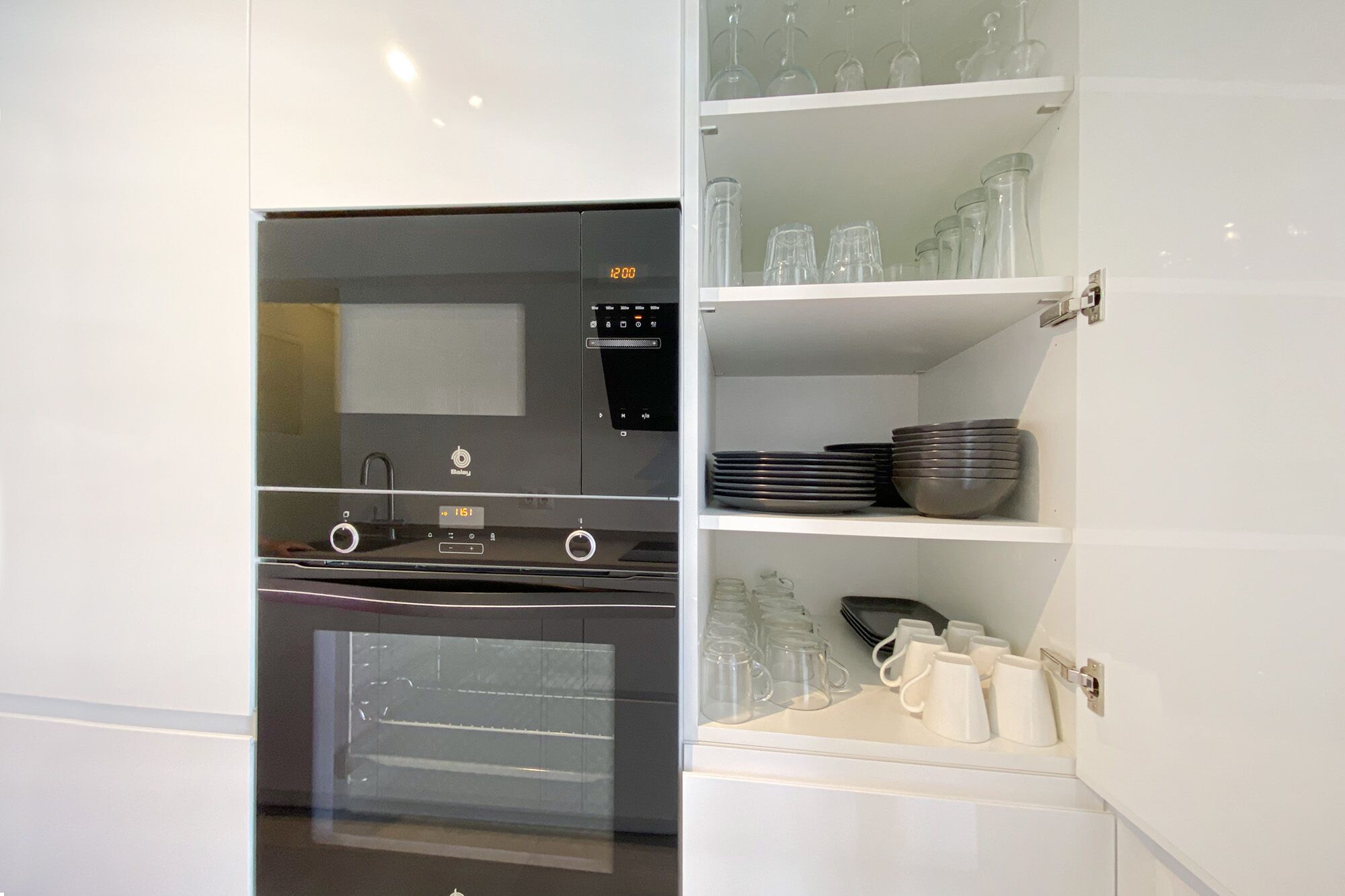 A modern kitchen with a built-in oven, microwave, and a glass-fronted cabinet with dishes and cups.