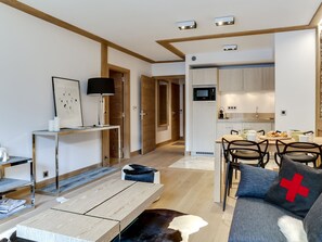 Uma sala de estar moderna com piso de madeira, área de jantar com mesa e cadeiras, cozinha com micro-ondas e um sofá com almofada vermelha.