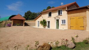 Una casa de dues plantes amb una porta verda, una porta de garatge de fusta i una petita zona coberta amb cadires.