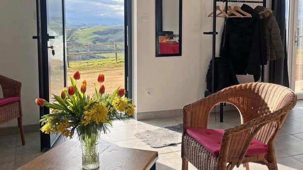Habitació amb vistes a un paisatge verd, una cadira de vimet, una taula amb un ram de flors i un penjador amb roba.