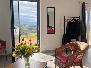 Ein Zimmer mit Blick auf eine grüne Landschaft, ein Korbstuhl, ein Tisch mit Blumen und ein Kleiderständer mit Kleidung.