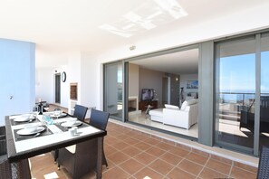 A balcony with a dining table set for four, a sofa, and a television in the background. - Apt with spacious terrace with amazing sea views, pool, 8 min drive to beach! (Mijas costa)