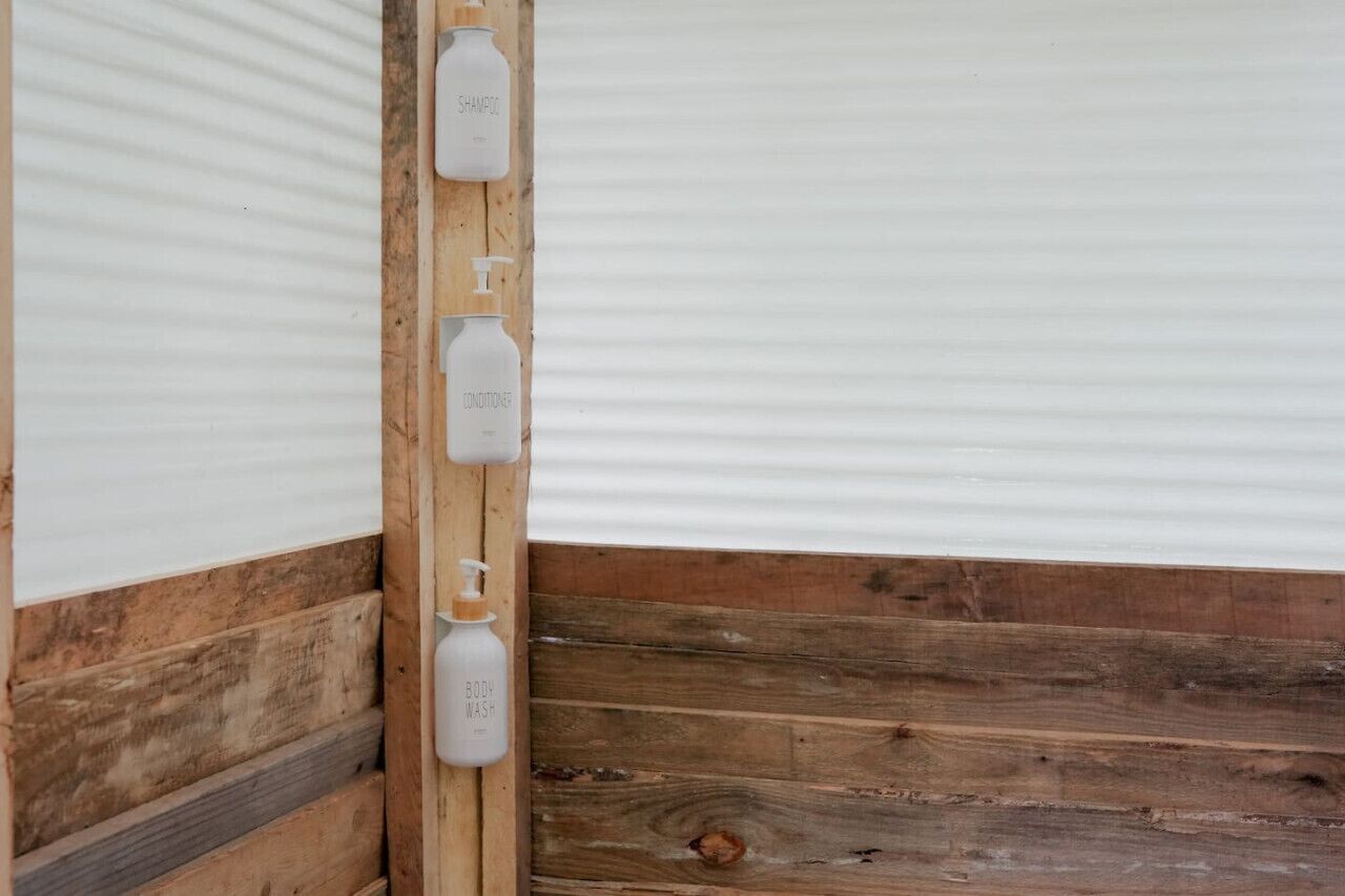 Een hoek van een kamer met houten lambrisering en een witte muur. Drie witte flesjes met de aanduiding 'SHAMPOO', 'CONDITIONER' en 'BODY WASH' zijn aan de muur bevestigd.