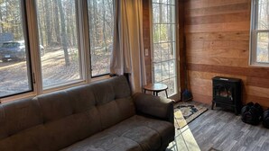 Interior acogedor de una cabaña con sofá, una mesita, chimenea y ventanas amplias con vistas al bosque circundante.