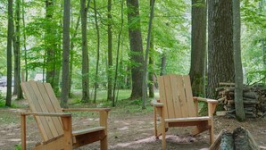 Dos sillas Adirondack de madera en un área boscosa, con un montón de leña cerca.