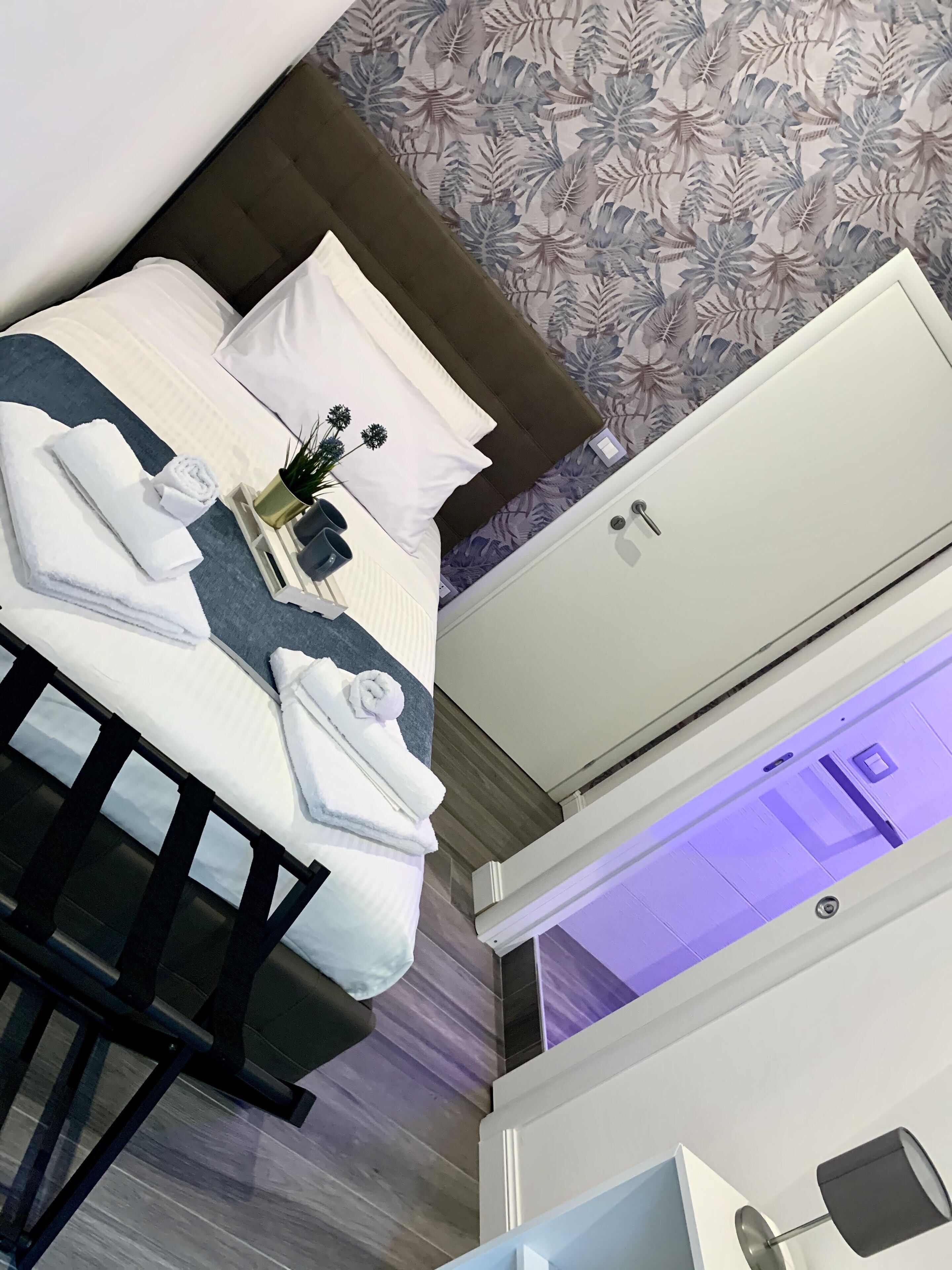 A hotel room with a bed, a white air conditioning unit, a wooden floor, and a floral wallpaper pattern.