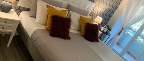 Un dormitorio con papel tapiz estampado, cabecera tapizada y cama con ropa de cama gris y dos almohadas rojas.
