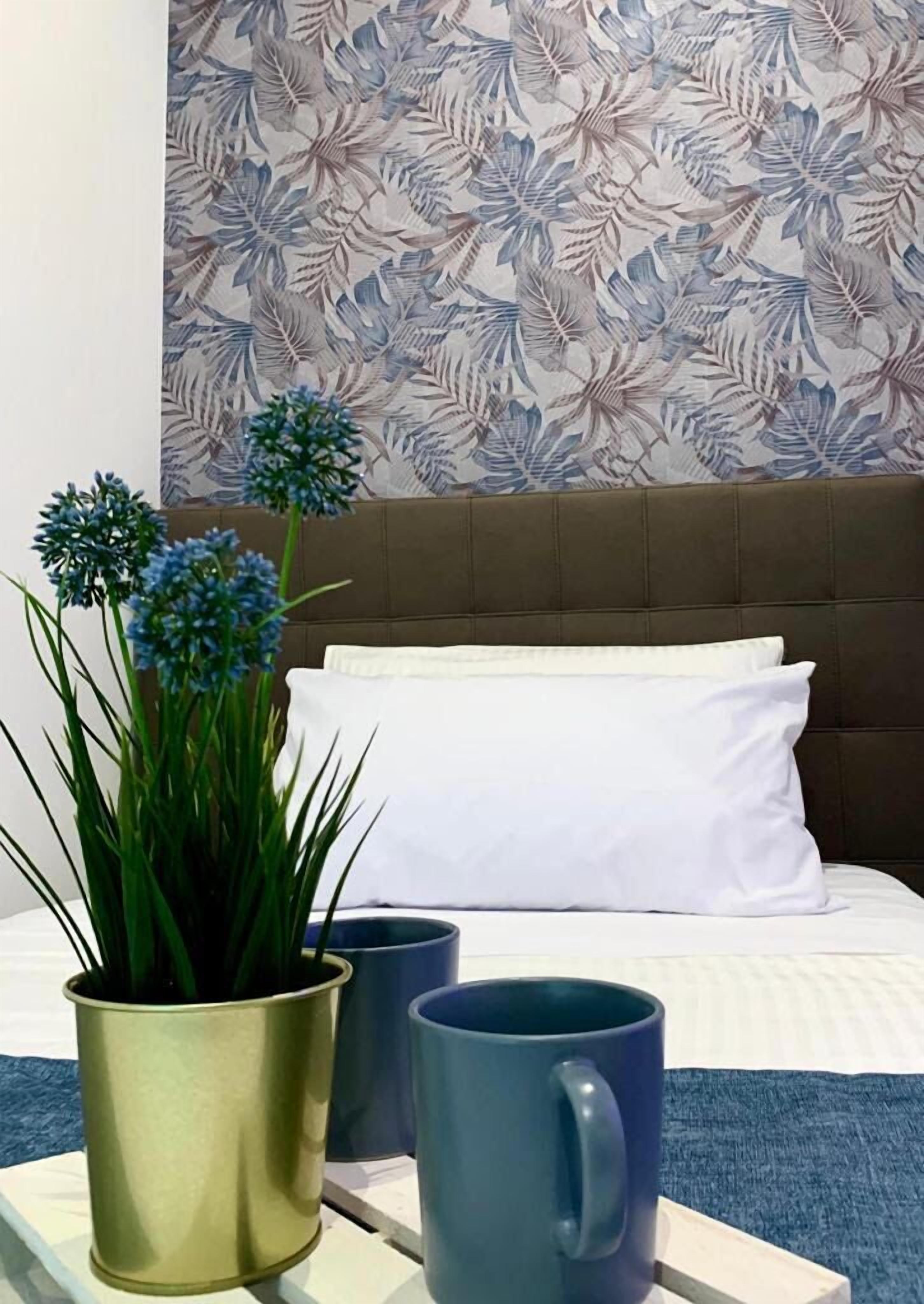 A hotel room with a bed, white pillows, a decorative wall, a potted plant, and two mugs.