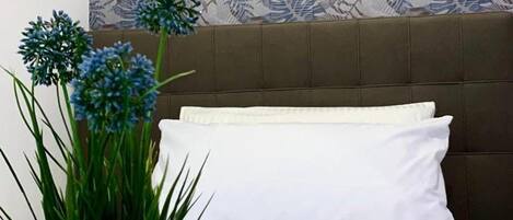 A hotel room with a bed, white pillows, a decorative wall, a potted plant, and two mugs.