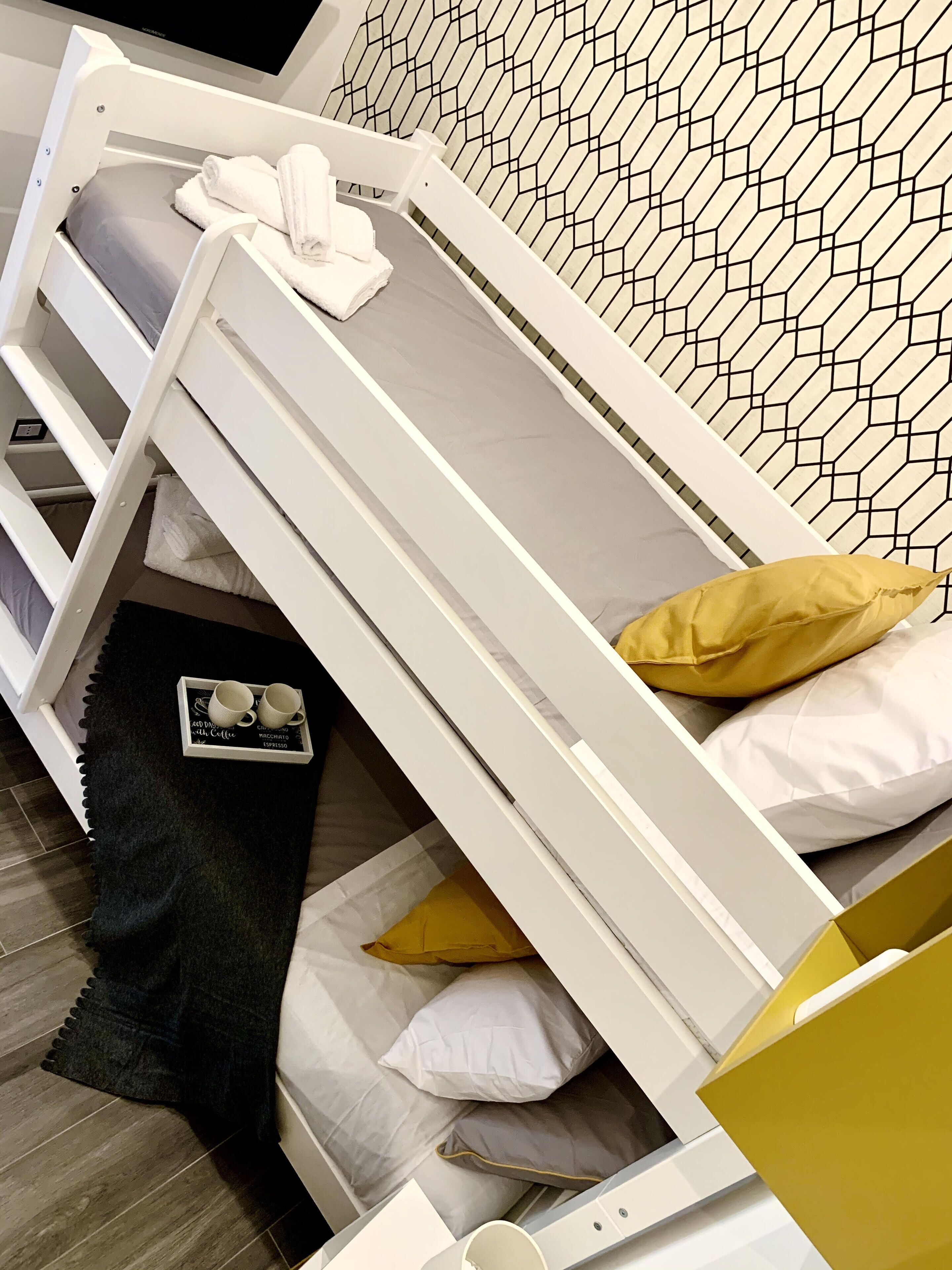 Una cama blanca con cabecera de diseño geométrico, una bandeja con dos tazas y una cuchara, y un almohadón amarillo.