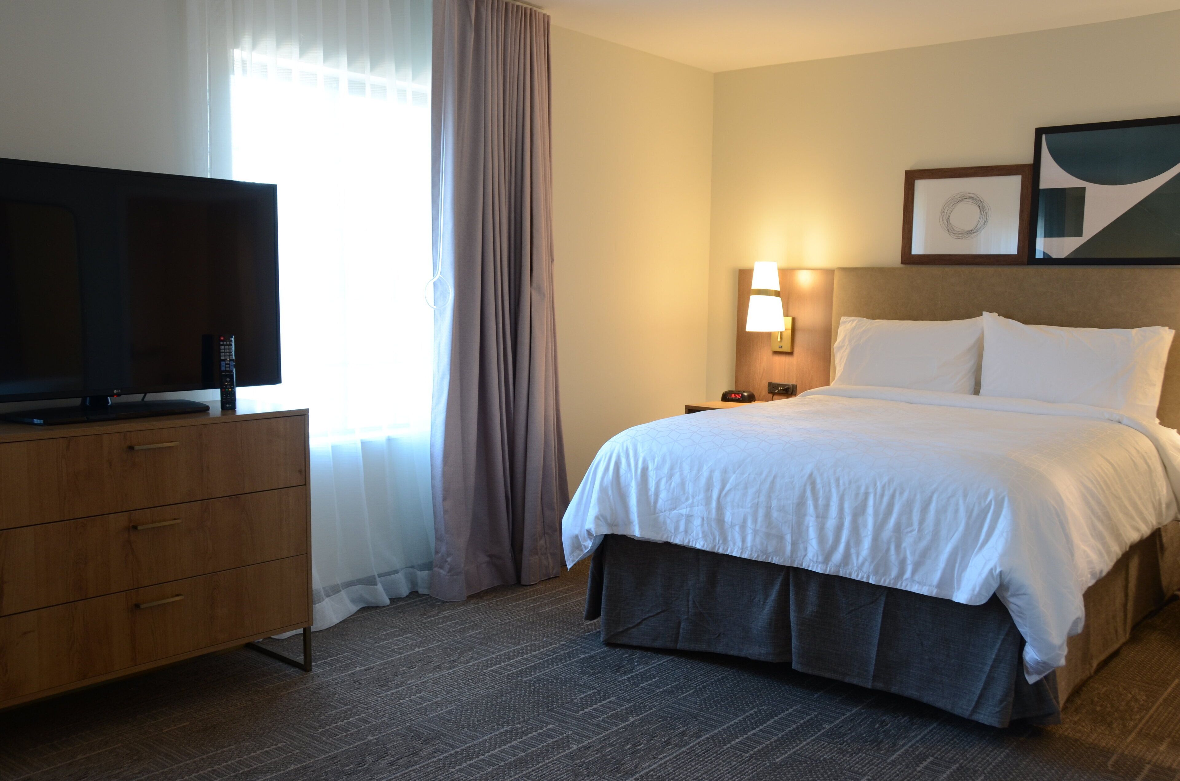 A hotel room with a bed, a television on a wooden dresser, a nightstand with a lamp, and a window with curtains.