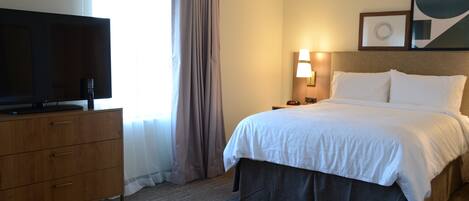 A hotel room with a bed, a television on a wooden dresser, a nightstand with a lamp, and a window with curtains.