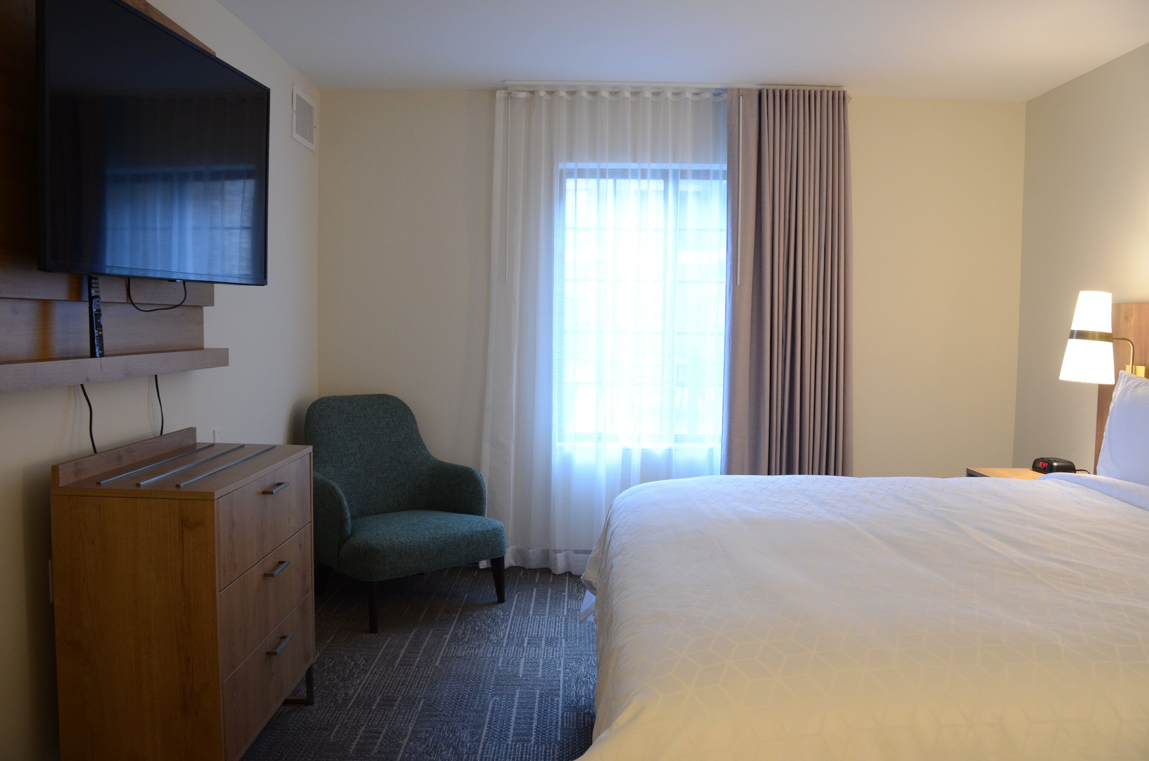 Ein Hotelzimmer mit einem Bett, einem Schreibtisch mit Fernseher, einem Sessel, einem Nachttisch mit Lampe und einem Fenster mit Vorhängen.