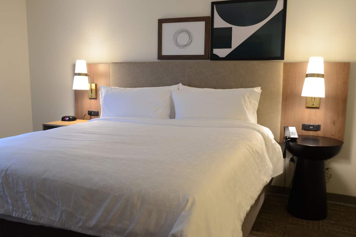 Una cama bien tendida con sábanas blancas, dos mesitas de noche con lámparas, un teléfono en la pared y una obra de arte abstracto sobre la cama.