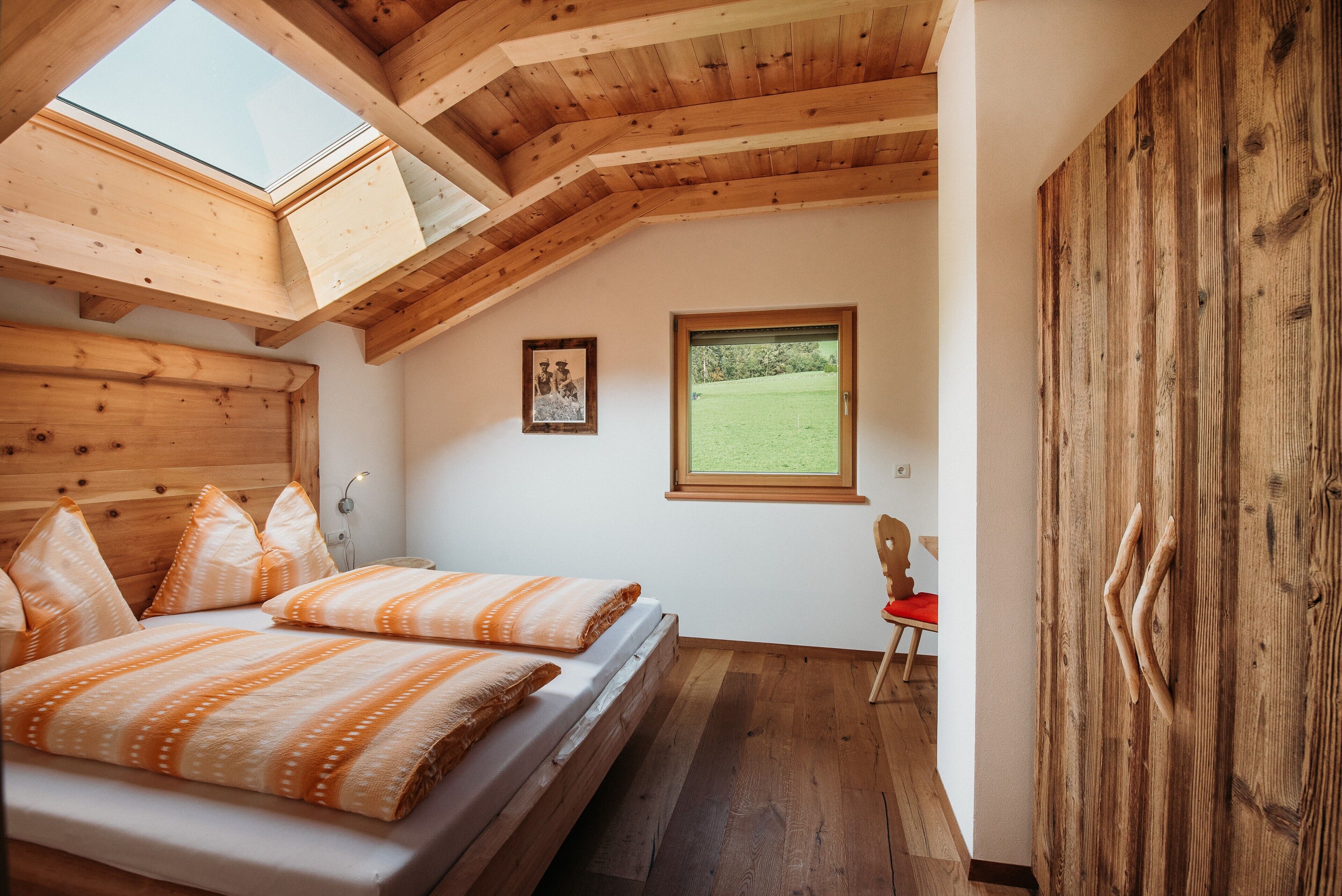 Een slaapkamer met een schuine plafond, houten balken, een bed met gestreept beddengoed, een ingelijst schilderij aan de muur, en een raam met uitzicht op een groen veld.