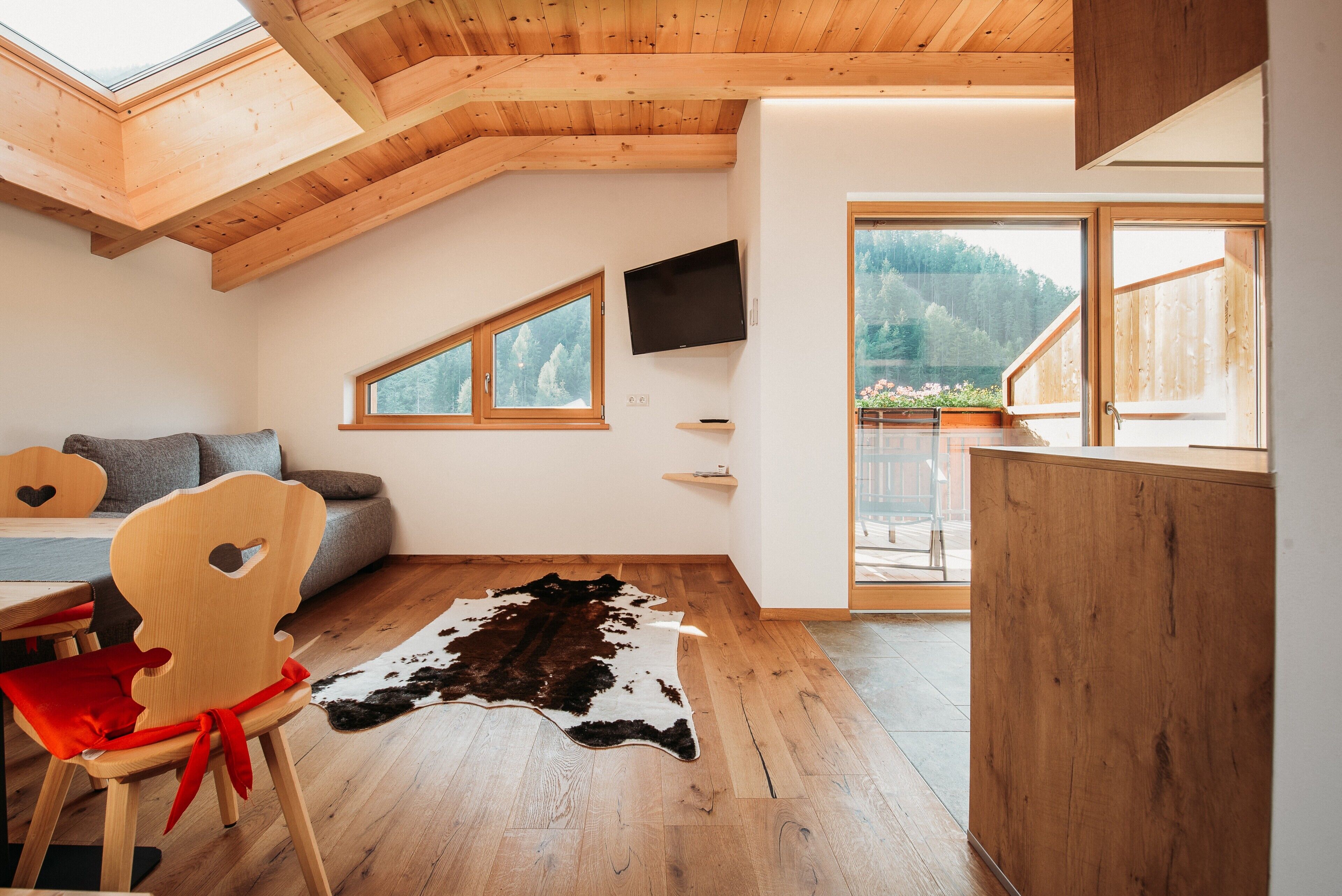 A room with a sloped wooden ceiling, a mounted TV, a large window with a view of trees, a wooden dining table, and a red chair.