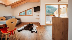 A room with a sloped wooden ceiling, a mounted TV, a large window with a view of trees, a wooden dining table, and a red chair. - Holiday Apartment “Lahnbach Großkarneid” in Beautiful Scenery with Mountain View (Lüsen)