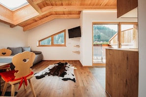 A room with a sloped wooden ceiling, a mounted TV, a large window with a view of trees, a wooden dining table, and a red chair.