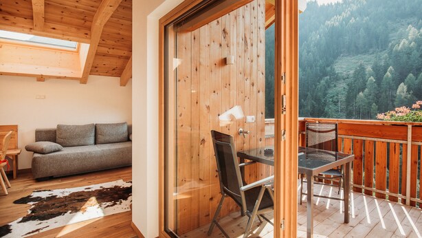 A wooden interior with a sofa, a dining table with chairs, and a glass door leading to a balcony with a view of a forested hillside. - Holiday Apartment “Lahnbach Großkarneid” in Beautiful Scenery with Mountain View (Lüsen)