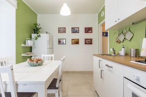 Una cocina con una mesa blanca, sillas, una lámpara colgante, un refrigerador y una pared con cuadros.