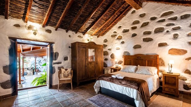 Un dormitorio rústico con pared de piedra, vigas de madera en el techo, una cama con colcha marrón, un armario de madera y una mesita de noche con lámpara.
