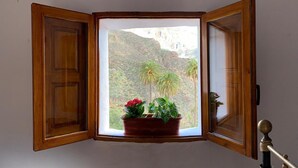 Una ventana en esquina con contraventanas de madera, una planta en maceta con flores rojas y vistas a un paisaje montañoso con palmeras.