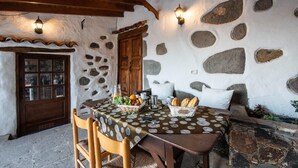 Una habitación rústica con paredes de piedra, una puerta de madera, una mesa dispuesta con una cesta de pan, frutas y una jarra, y dos luces fijadas a la pared.