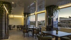 An indoor dining area with tables and chairs, large windows, and hanging plants. - Royalty Hotel Athens (Athens)