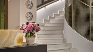 A reception area with a marble counter, a vase of flowers, two wall clocks, and a staircase with a circular glass railing. - Royalty Hotel Athens (Athens)
