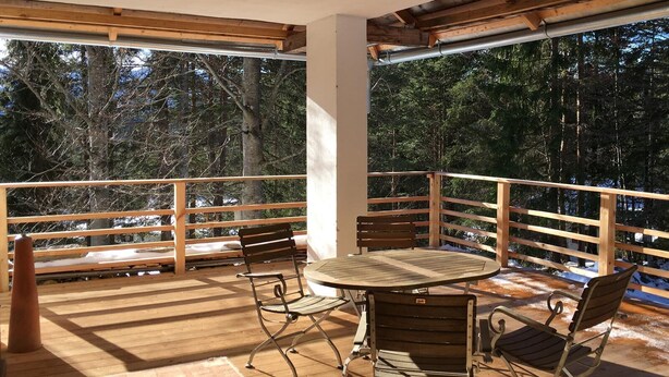 Una terraza de madera con una mesa redonda y sillas metálicas, rodeada por un barandal de madera, con vistas a un área boscosa.