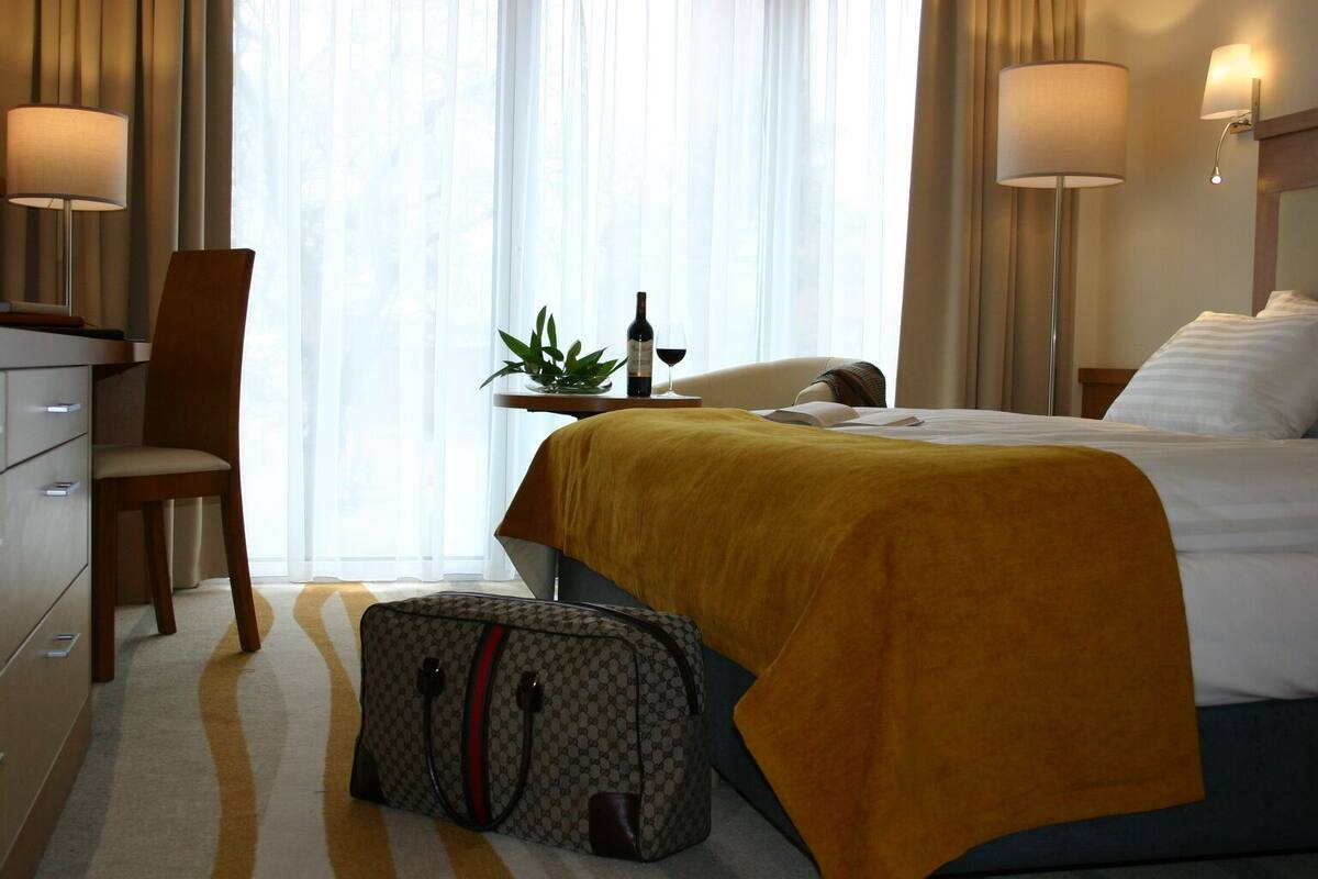 Ein Hotelzimmer mit Bett, Schreibtisch, Stuhl, Lampe, Pflanze und Koffer.