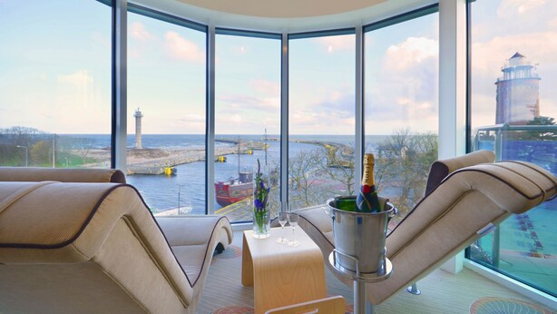 A corner room with a large window offering a view of a body of water, a lighthouse, and a bridge. There is a cushioned seating area with a table holding a bottle of champagne and two glasses. - Hotel Hanseatic  - adults only (Kolobrzeg)