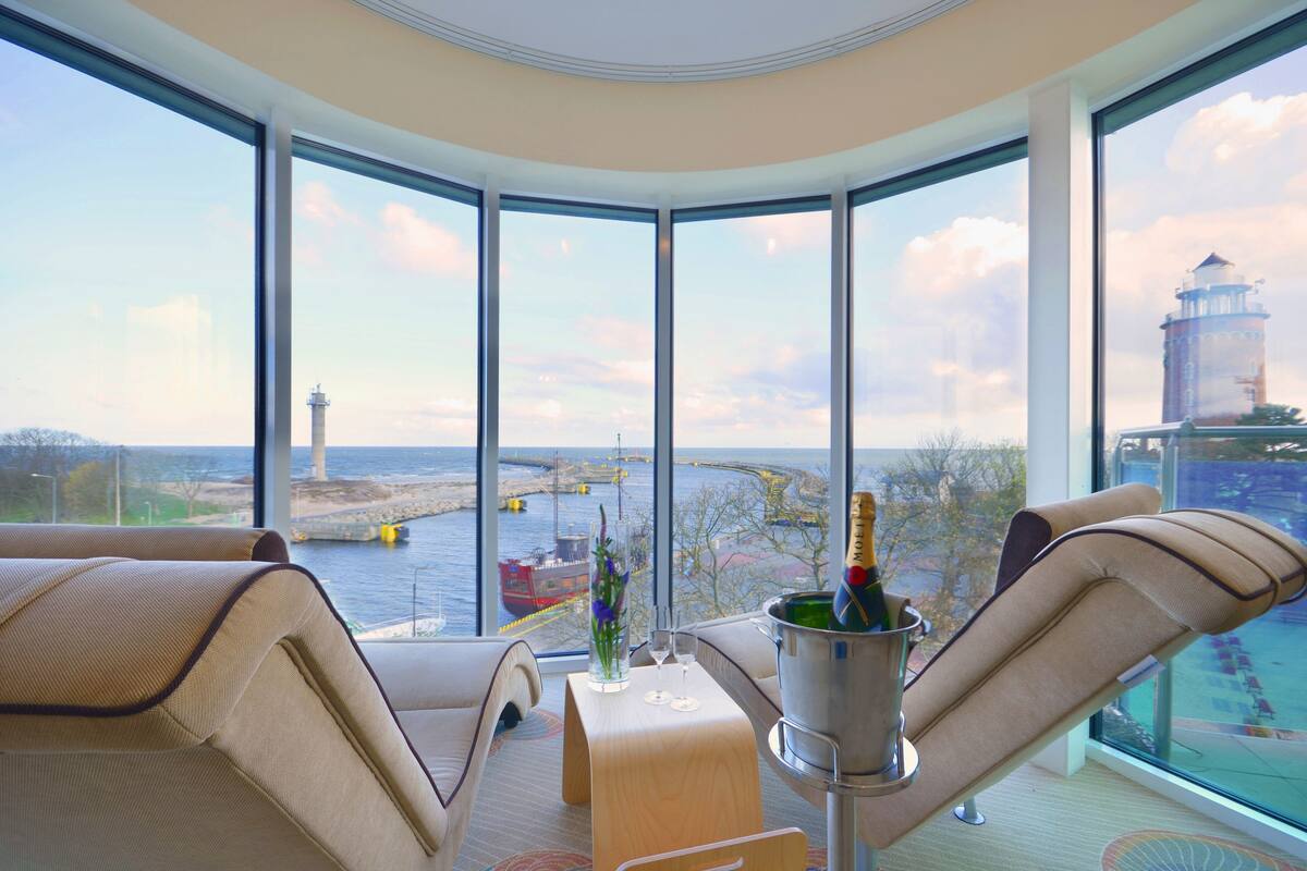 Ein Eckzimmer mit großem Fenster und Blick auf ein Gewässer, einen Leuchtturm und eine Brücke. Es gibt eine gepolsterte Sitzecke mit einem Tisch, auf dem eine Champagnerflasche und zwei Gläser stehen.
