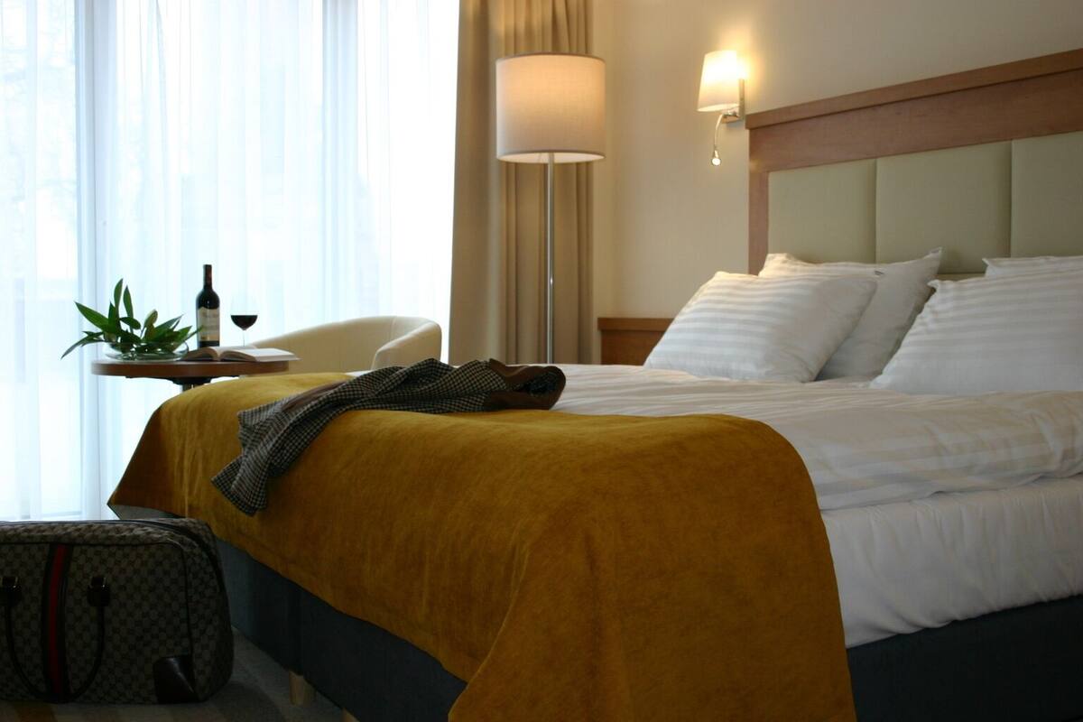 Ein Hotelzimmer mit Bett, einer Nachttischlampe, einem Tisch mit Pflanze und Flasche sowie einem Koffer.