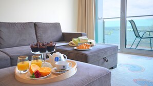 A modern living room with a grey sofa, a dining table with breakfast items, and a view of the sea through large windows. - Hotel Hanseatic  - adults only (Kolobrzeg)