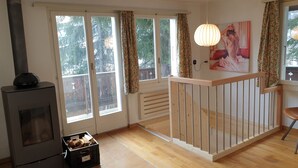 Une pièce chaleureuse avec un plancher en bois, une cheminée, une balustrade en bois et de grandes fenêtres ornées de rideaux à motifs floraux.
