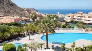 A resort with a large swimming pool surrounded by palm trees, a view of the sea, and Mediterranean-style buildings.