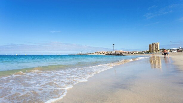 A beach with clear blue water, a sandy shore, and buildings in the background.