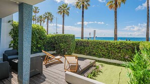 Un espace extérieur abrité, aménagé avec des meubles en osier, une terrasse en bois et offrant une vue sur la mer et les palmiers.