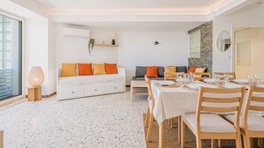 Un salon moderne avec un coin repas, des canapés blancs agrémentés de coussins orange, une cheminée et une grande fenêtre.