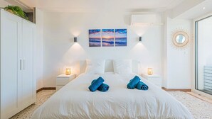 Une chambre à coucher comprenant un lit, deux tables de chevet, un climatiseur fixé au mur, une grande fenêtre et un miroir décoratif.