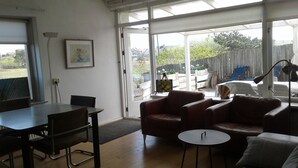 Ein Wohnzimmer mit Esstisch, zwei Sofas und einem kleinen runden Tisch. Ein Gemälde hängt an der Wand und ein Fenster bietet Blick auf den Garten.