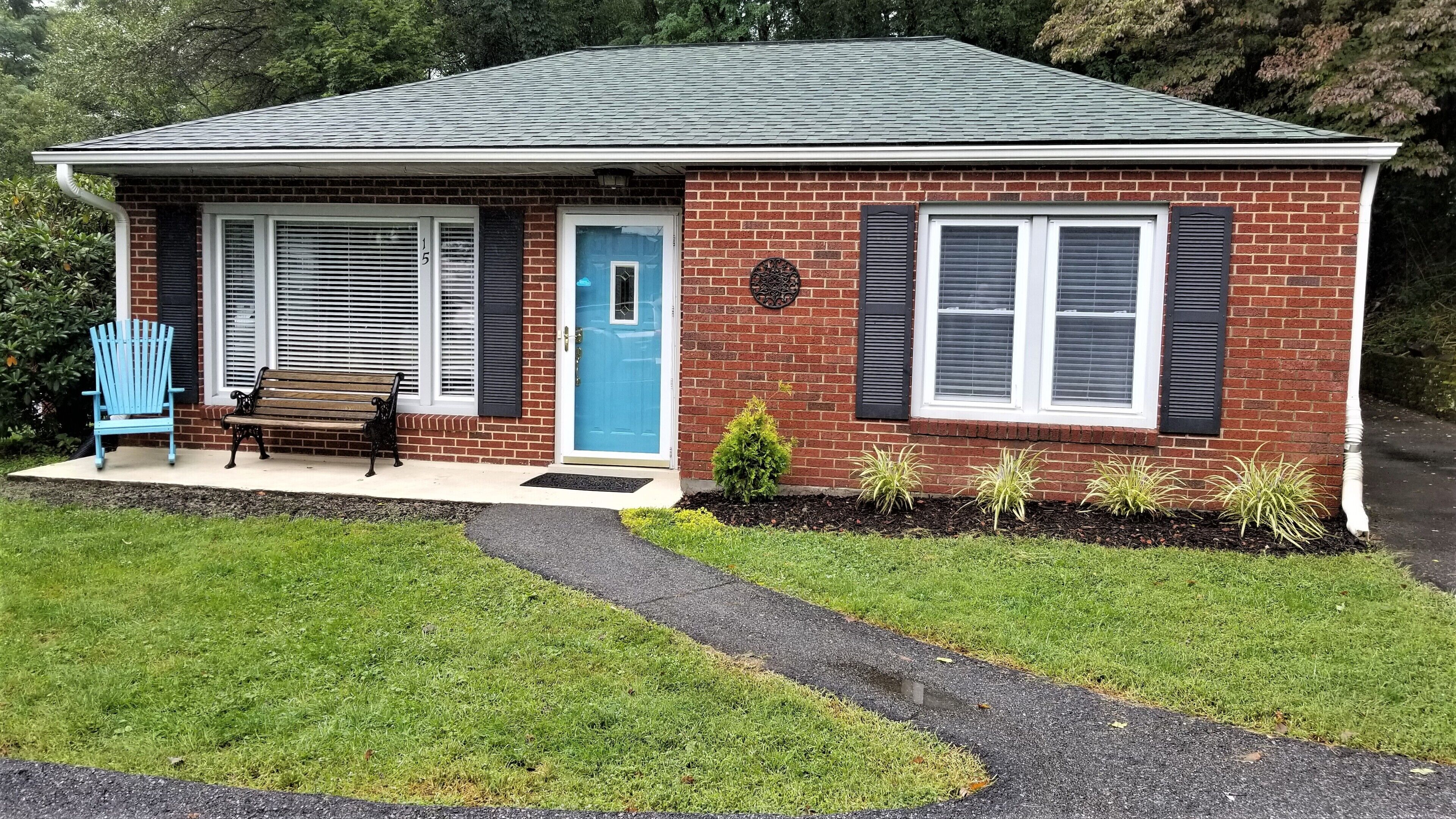 A single-story brick house with a blue front door, two windows with shutters, a small porch with a bench, and a well-maintained lawn.