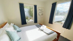 A neatly made bed with pillows and a towel, two windows with curtains, and a view of a mountain landscape. - RIMA | Sun-Soaked Hideaway (Queenstown)