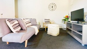 A living room with a grey sofa, a white ottoman, a wooden chair, a TV stand, and a wall clock. - RIMA | Sun-Soaked Hideaway (Queenstown)