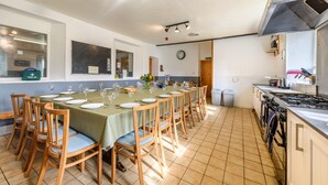 Ein Essbereich mit einem langen Tisch, gedeckt für eine Mahlzeit, Holzsessel, eine Küchenzeile mit Herd und Schränken sowie eine Uhr an der Wand.